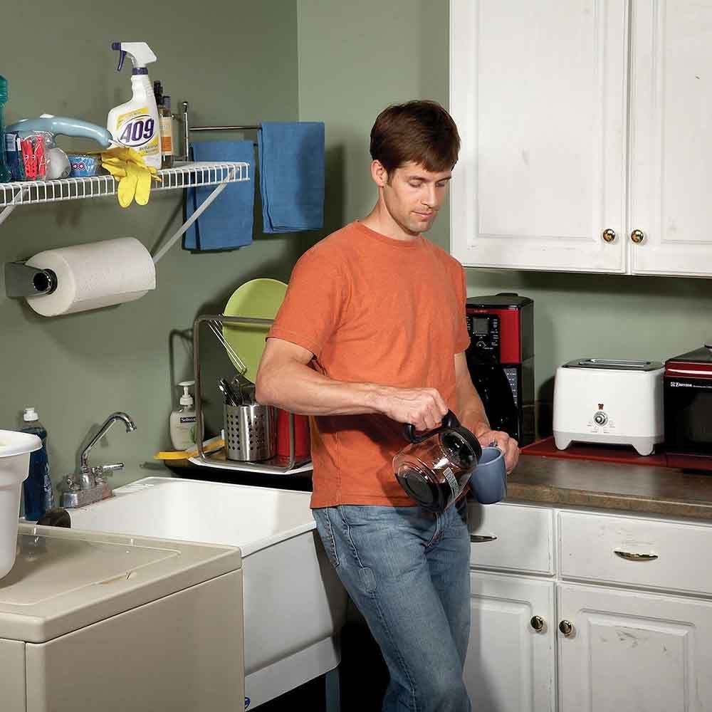 A man in an orange shirt pours coffee from a carafe into a mug in a kitchen with various appliances and cleaning supplies nearby.