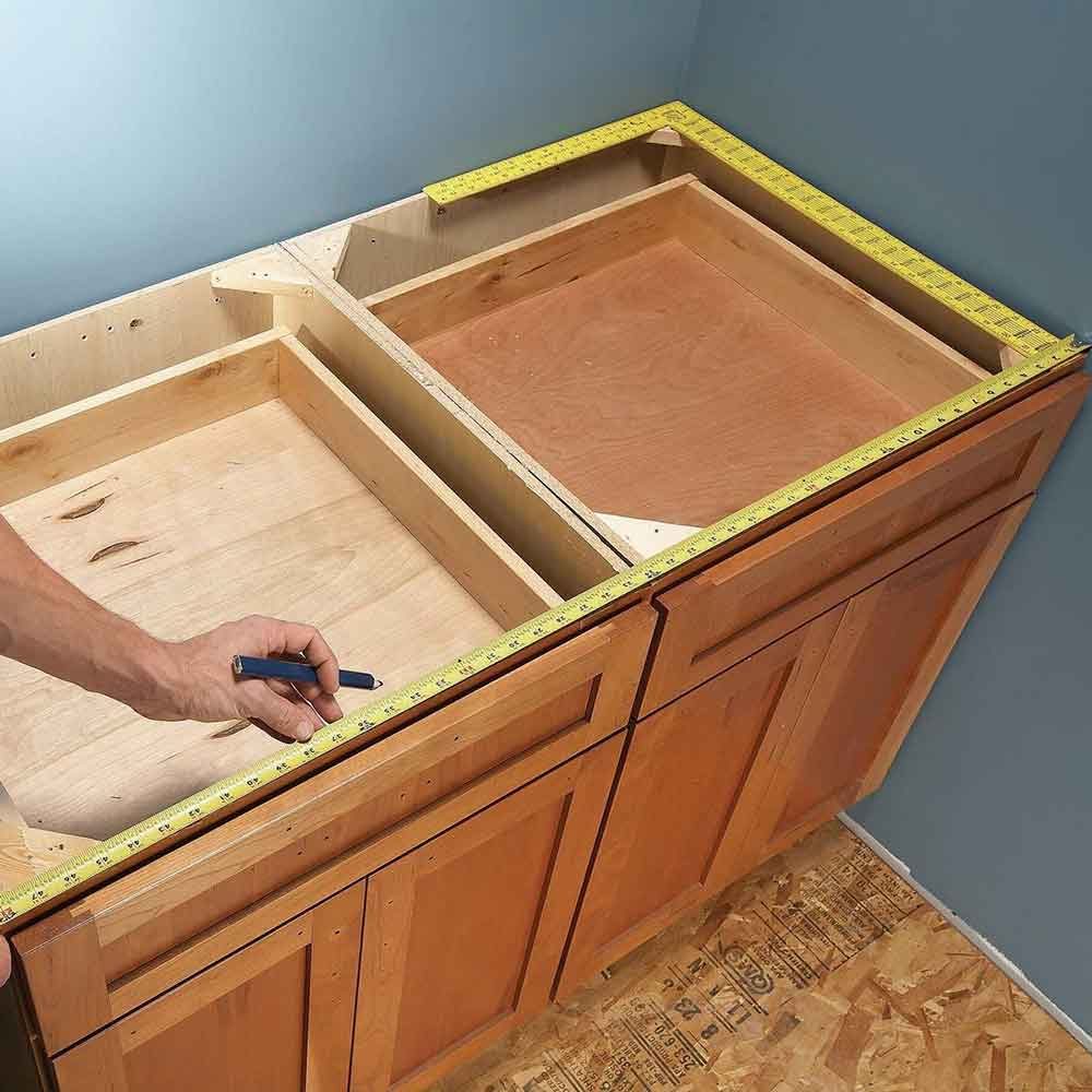 A hand measures a wooden cabinet surface with a tape measure, displaying an unfinished interior, set against a blue wall and a plywood floor.