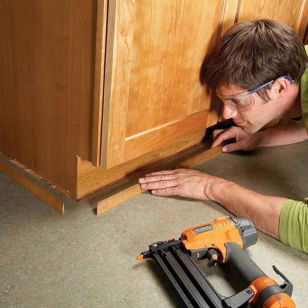 A man kneels, applying trim to a cabinet's base with a nail gun on a textured floor in a well-lit kitchen environment.