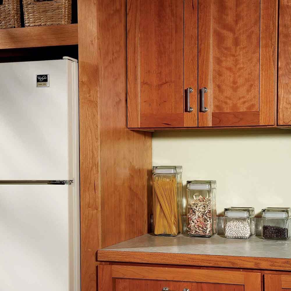 A white refrigerator stands near wooden cabinets. Glass jars filled with pasta and various beans are placed on a countertop against a light green wall.