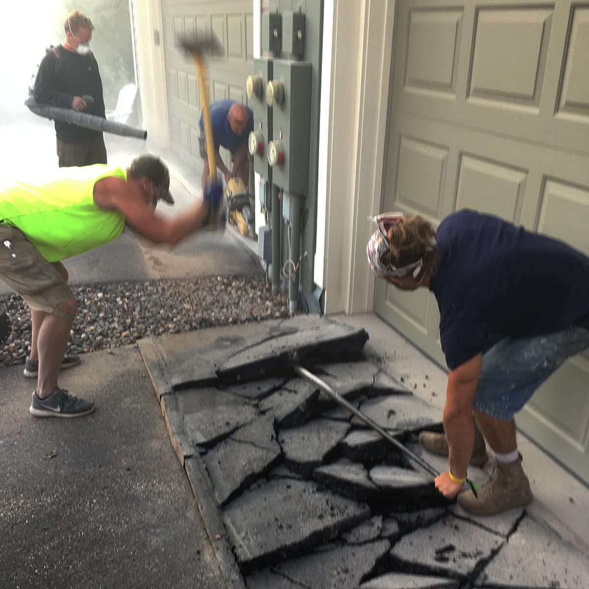 Workers are breaking apart cracked pavement outside a garage, using a hammer and pry bar, with dust in the air and electrical panels nearby.