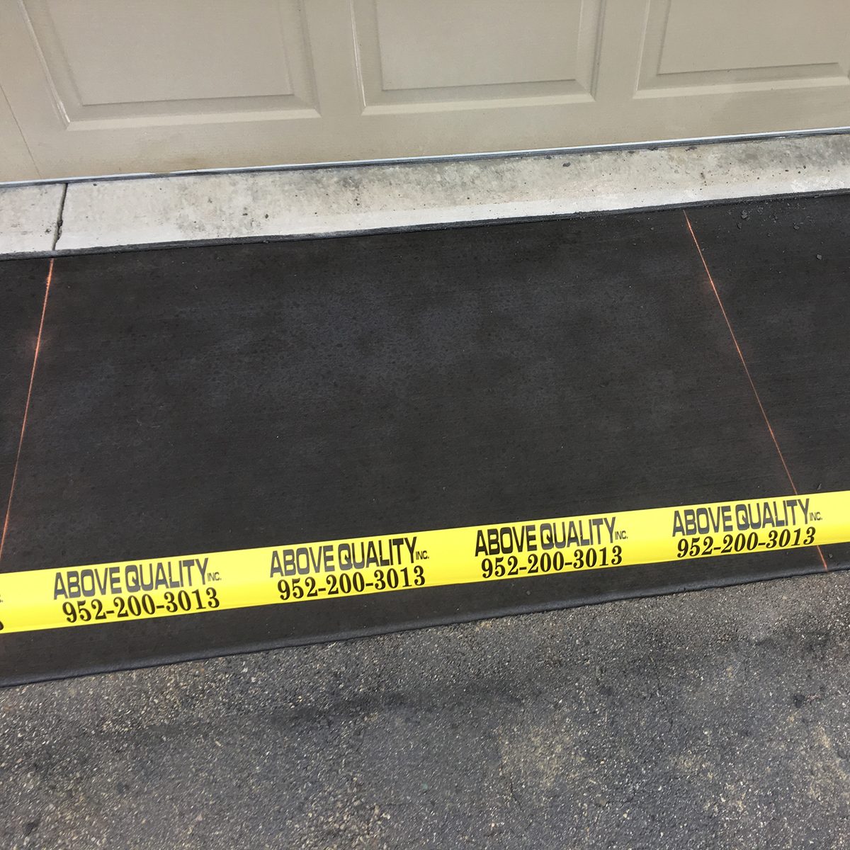 A yellow banner reading "ABOVE QUALITY, inc. 952-200-3013" rests on a dark surface near a garage door, indicating potential work or caution in the area.