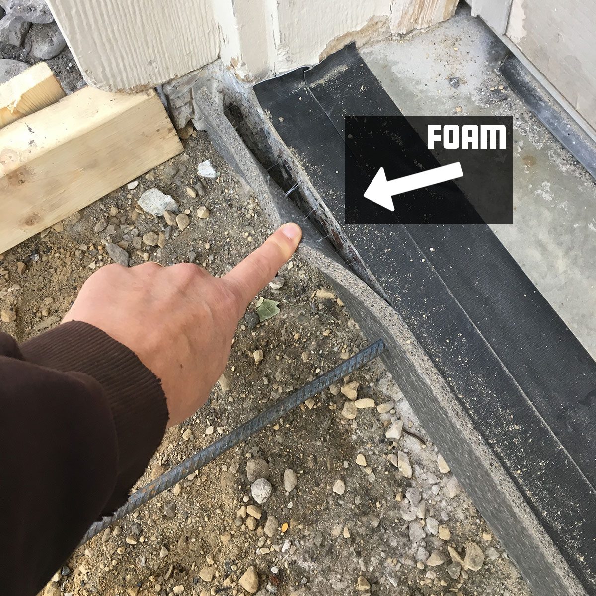 A finger points at a gap between a concrete surface and a wall, indicating where foam needs to be applied, surrounded by dirt and building materials.