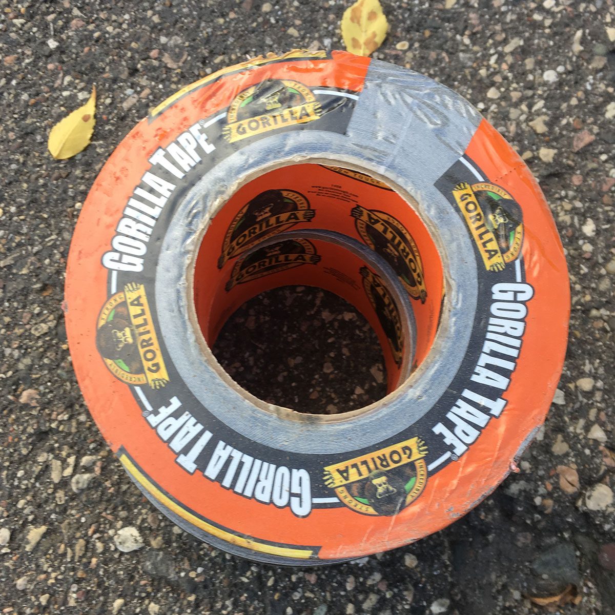 A roll of orange Gorilla Tape lies on a textured surface, showcasing its logo and partially visible inner layer, surrounded by scattered autumn leaves.