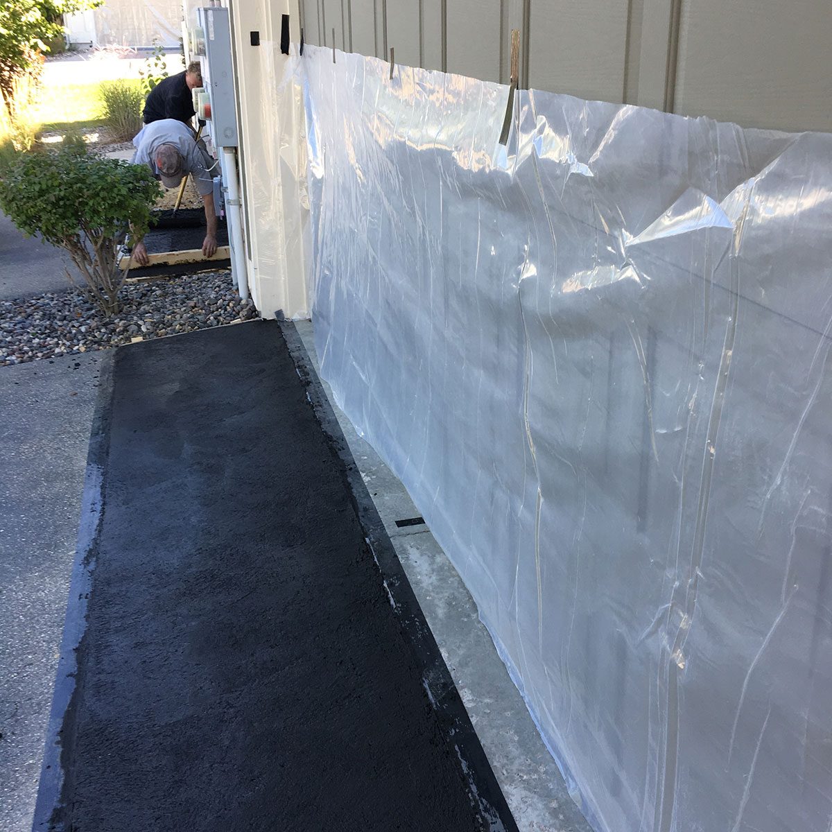 Two men are applying fresh black concrete along a wall, with plastic sheeting protecting adjacent areas. A shrub and pebbles are visible nearby.