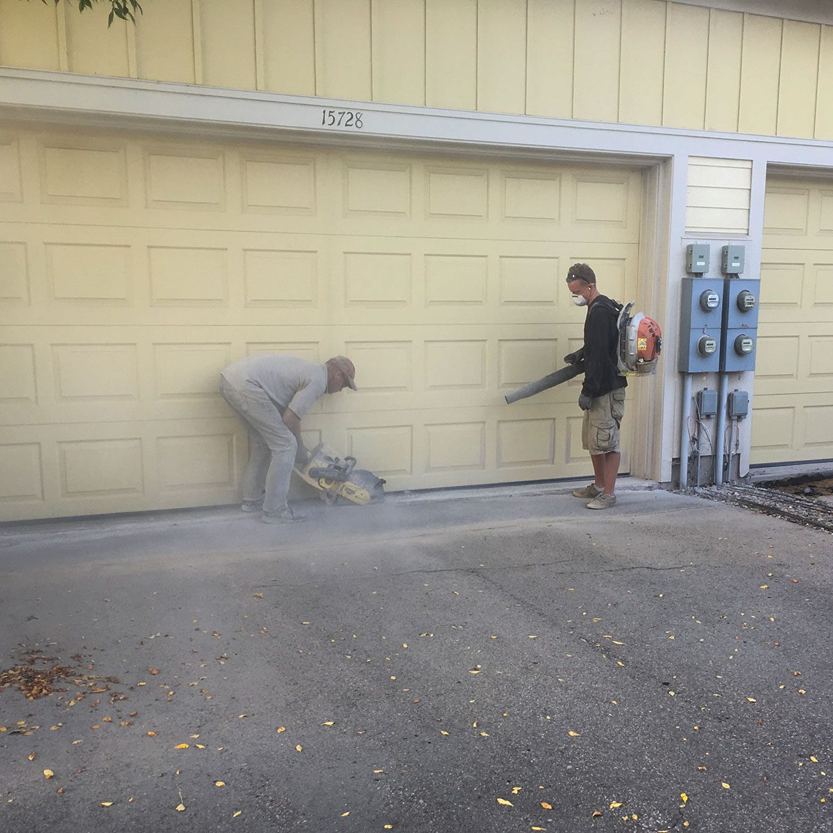A man cuts concrete near a garage door while another uses a leaf blower to disperse dust in a residential driveway setting.