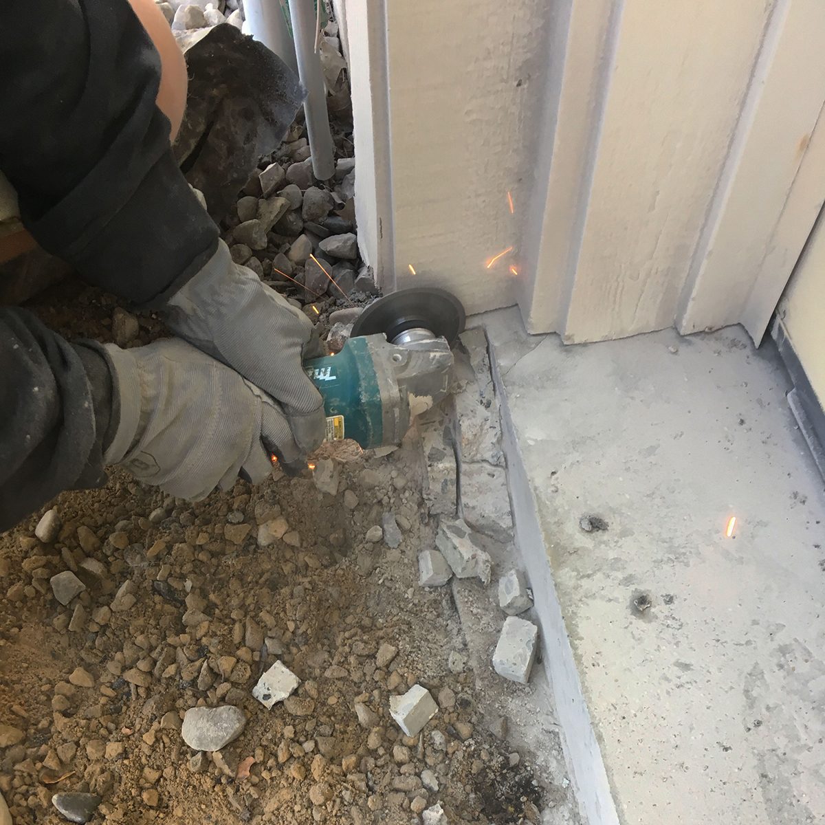 A person uses a power tool to cut concrete near a building corner, producing sparks, with dirt and small stones scattered around the base.