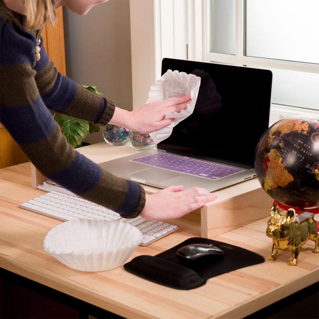 A person wipes a laptop screen with a coffee filter on a wooden desk. Nearby, a globe, mouse, and colorful decorations are visible.