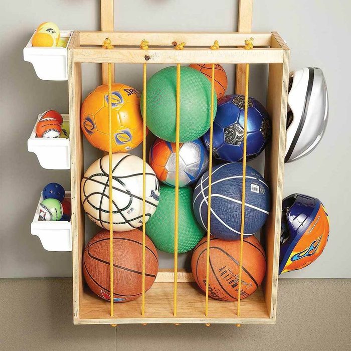 A wooden rack holds various sports balls and helmets, organized behind elastic bands, against a neutral-colored wall, conveying a neat sports storage solution.
