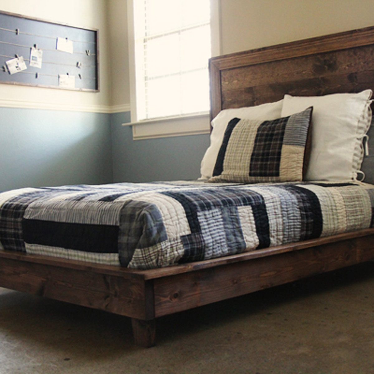 A wooden bed features a checkered quilt and pillows. Sunlight streams through a window, illuminating a simple, cozy bedroom with framed photos on the wall.