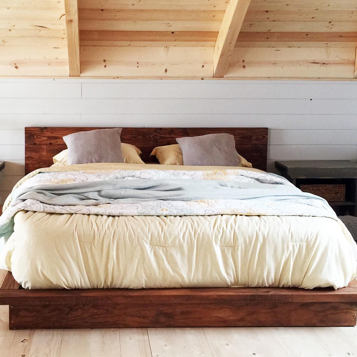 A wooden bed with a light yellow comforter and pillows is centered in a cozy room, featuring sloped wooden ceilings and a simple bedside table.
