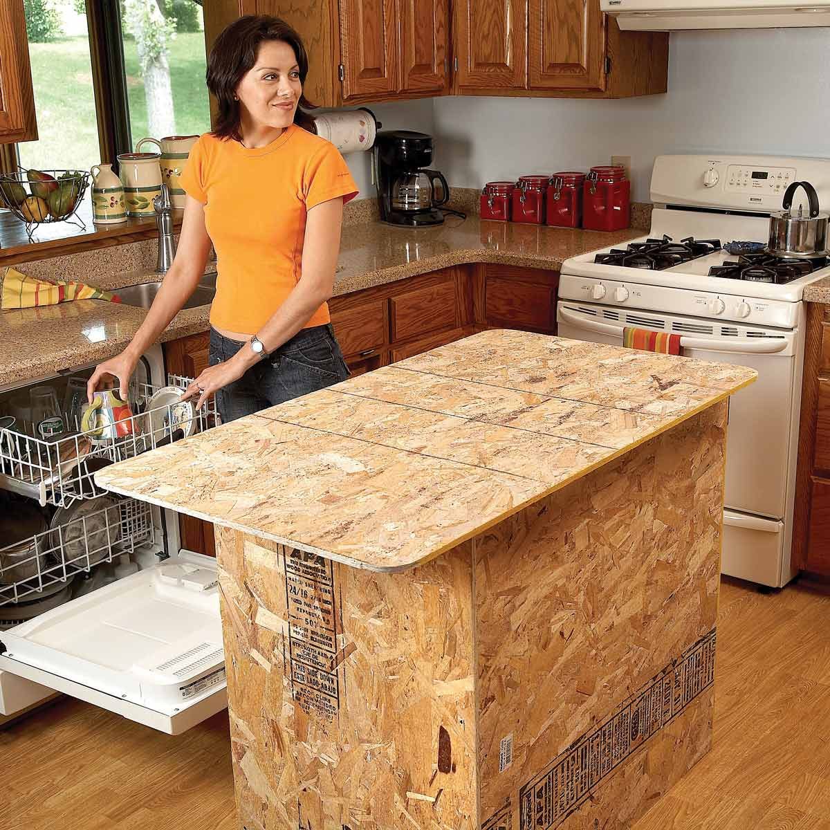 A woman unloads clean dishes from a dishwasher in a kitchen, featuring wooden cabinets, a countertop, and an unfinished island made of oriented strand board.