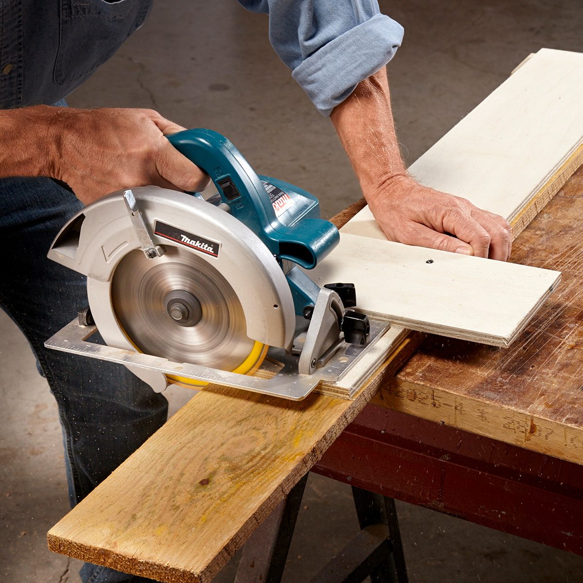 A circular saw cuts through a wooden plank, held steady by a person's hand, on a workbench in a workshop environment.