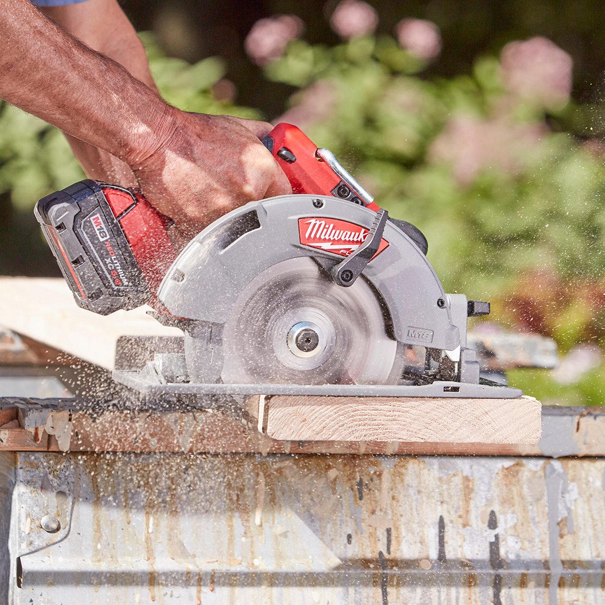 A person operates a circular saw, cutting through a wooden board, while wood shavings fly in the air. The background features blurred greenery.