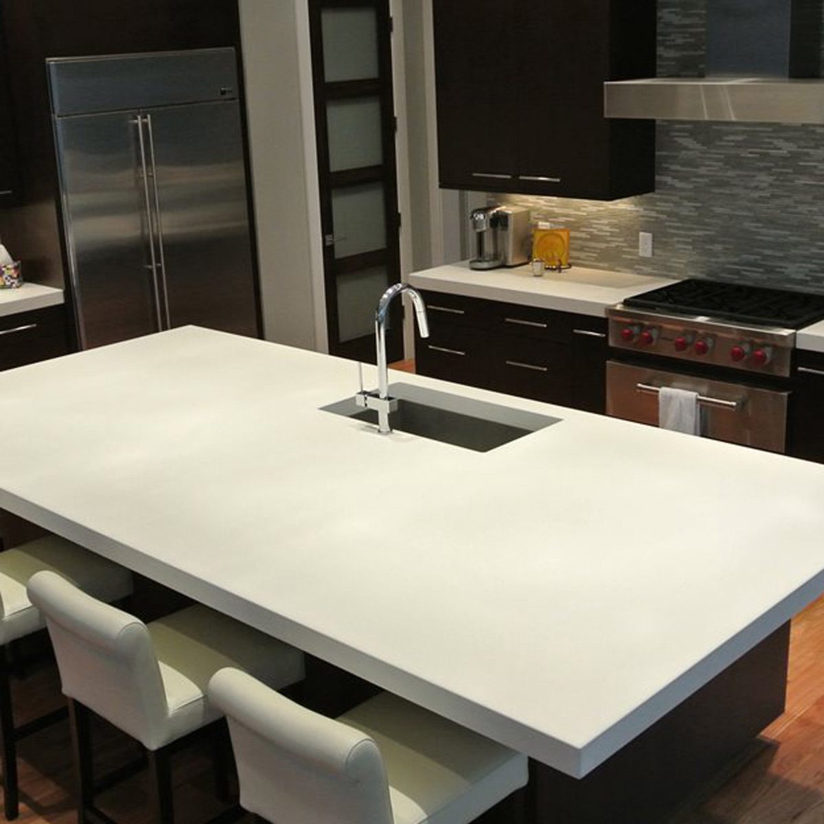 A large white kitchen island with a sink creates a central workspace, surrounded by modern dark cabinetry and stainless steel appliances in a contemporary setting.