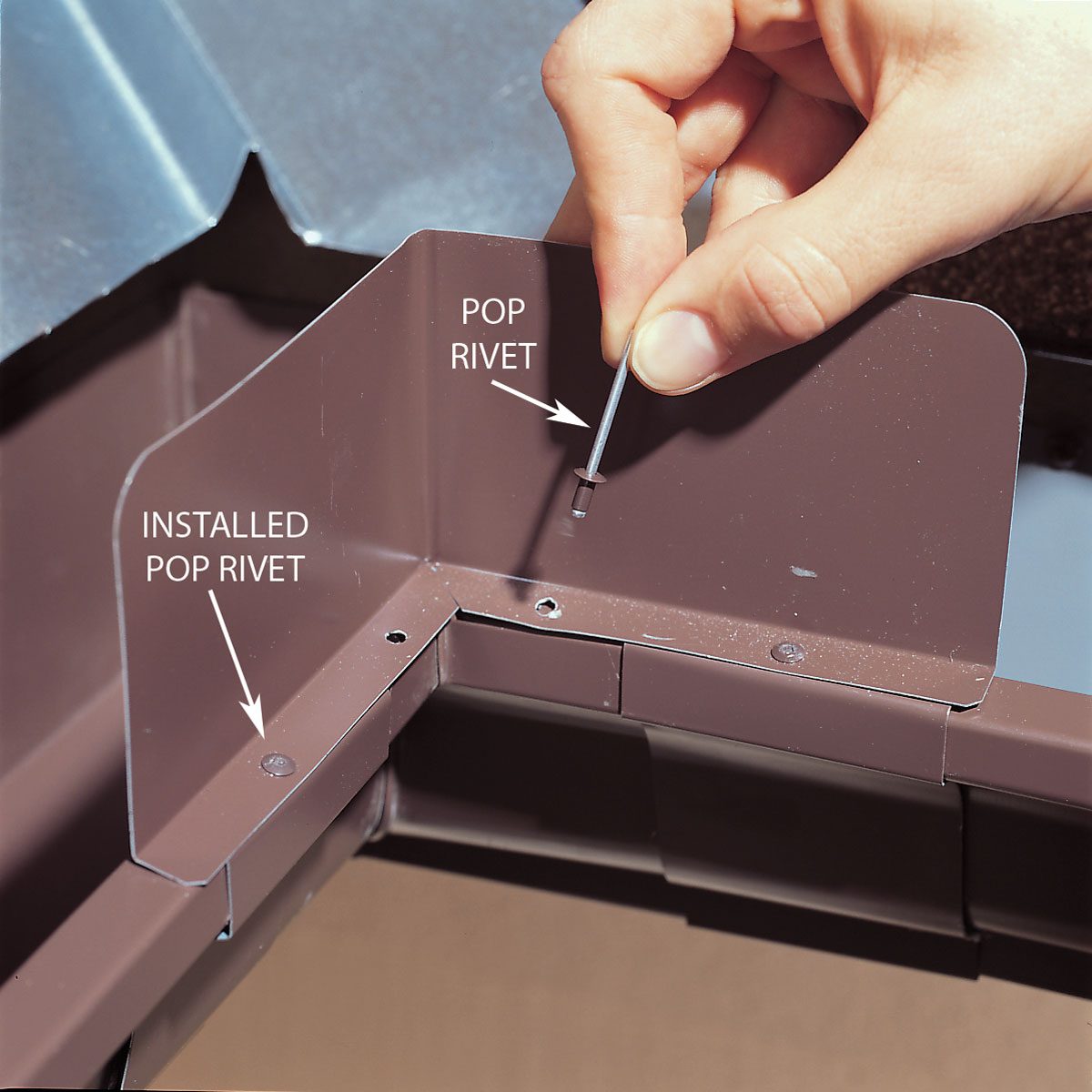 A hand inserts a pop rivet into a brown metal joint, demonstrating the installation process in a construction or repair setting.