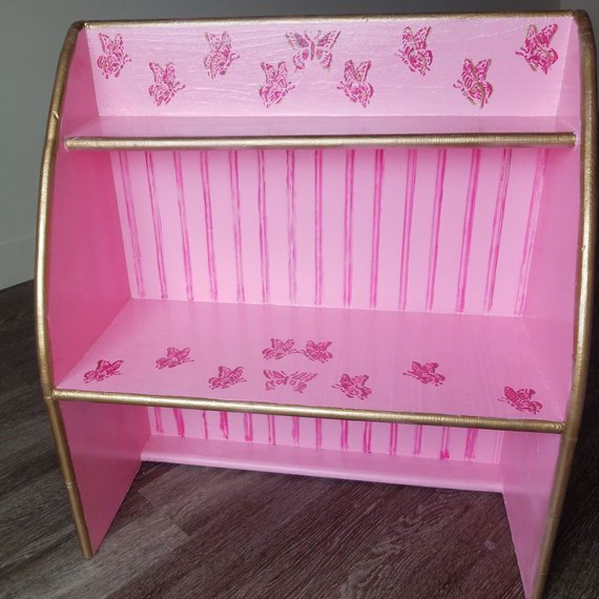 A wooden pink shelf with butterfly designs rests on a wooden floor, featuring two levels for storage and gold trim along the edges.