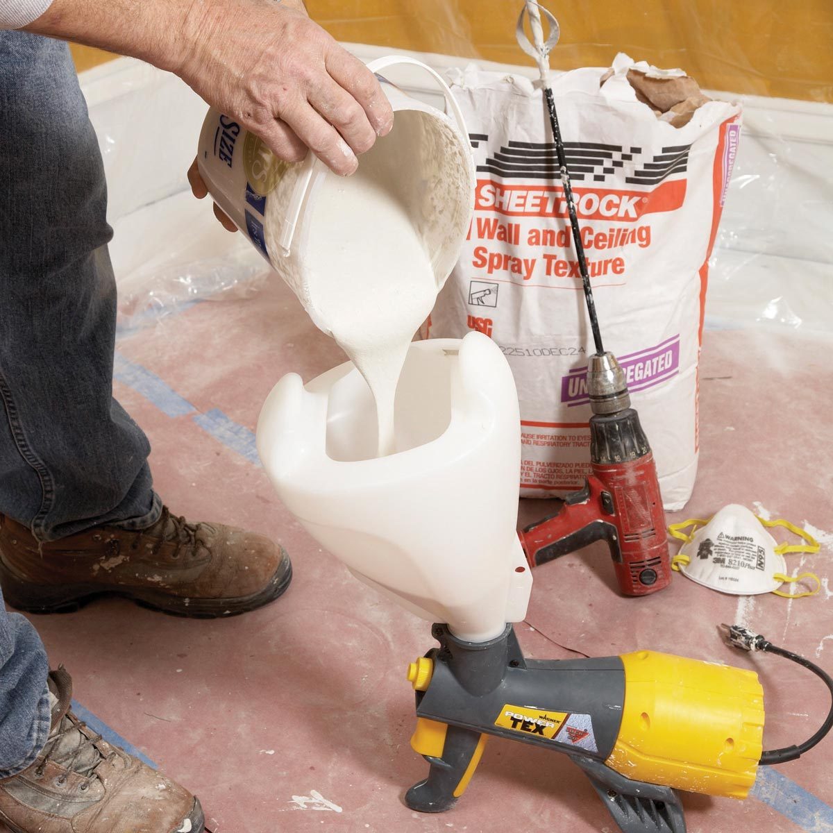 A person pours a light-colored substance from a bucket into a sprayer on the floor, surrounded by construction materials and tools.