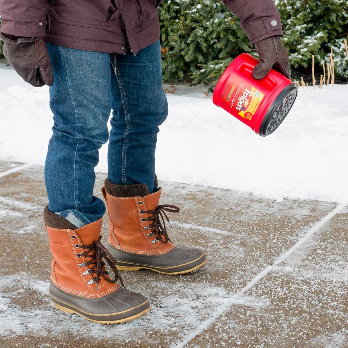 A person in winter boots holds a red container, spreading a substance on a snowy sidewalk while standing in a snowy outdoor environment.