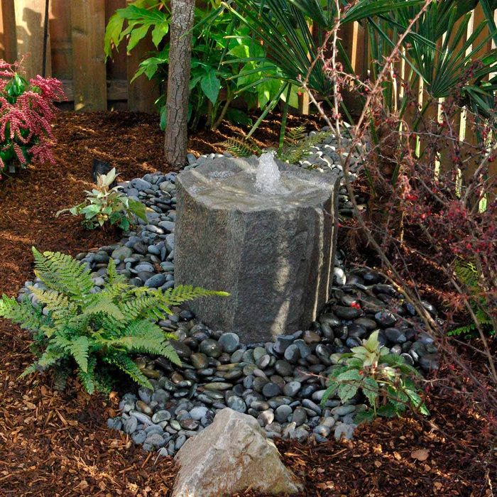 A stone fountain bubbles water, surrounded by dark pebbles and lush green plants in a serene garden setting, with wooden fencing in the background.