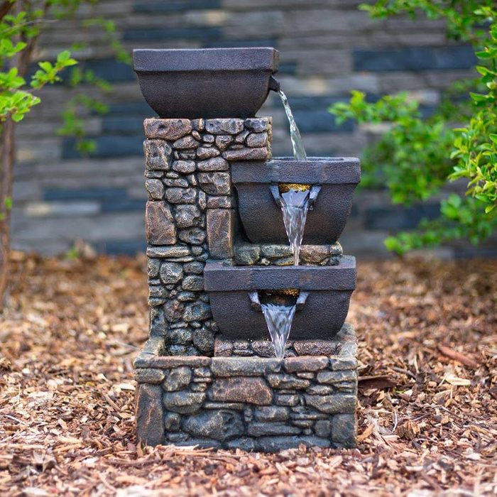 A stone fountain displays water cascading from three rectangular bowls, surrounded by mulch and greenery against a textured, dark background.