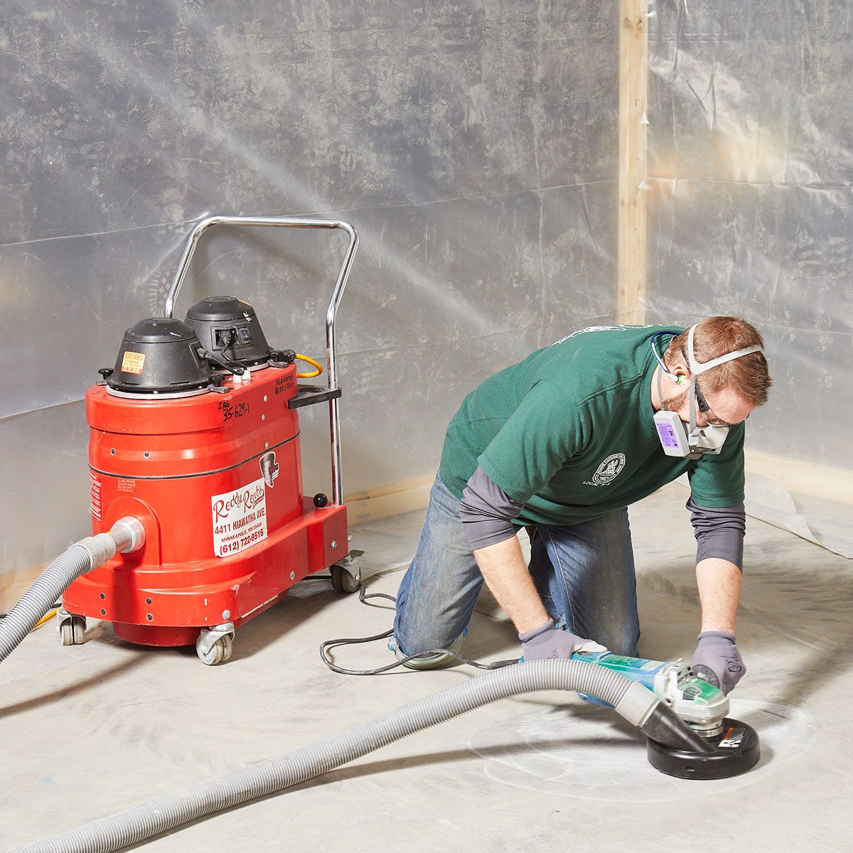 A man kneels on a concrete floor, operating a floor sanding tool connected to a red vacuum cleaner, in a partially enclosed, plastic-covered environment.