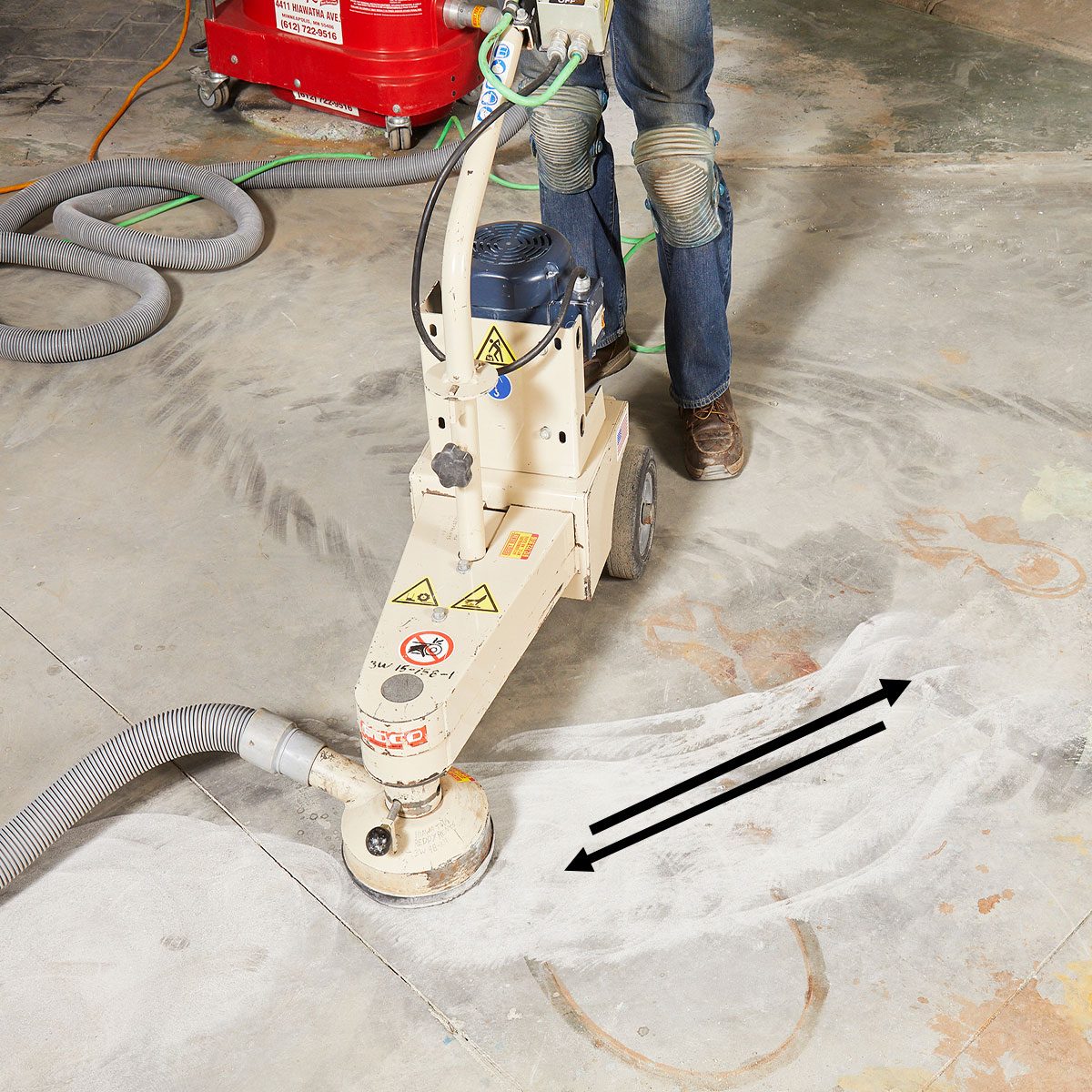 A floor grinding machine operates, smoothing a concrete surface while connected to a vacuum hose. Environmental clutter includes cords and markings on the floor.
