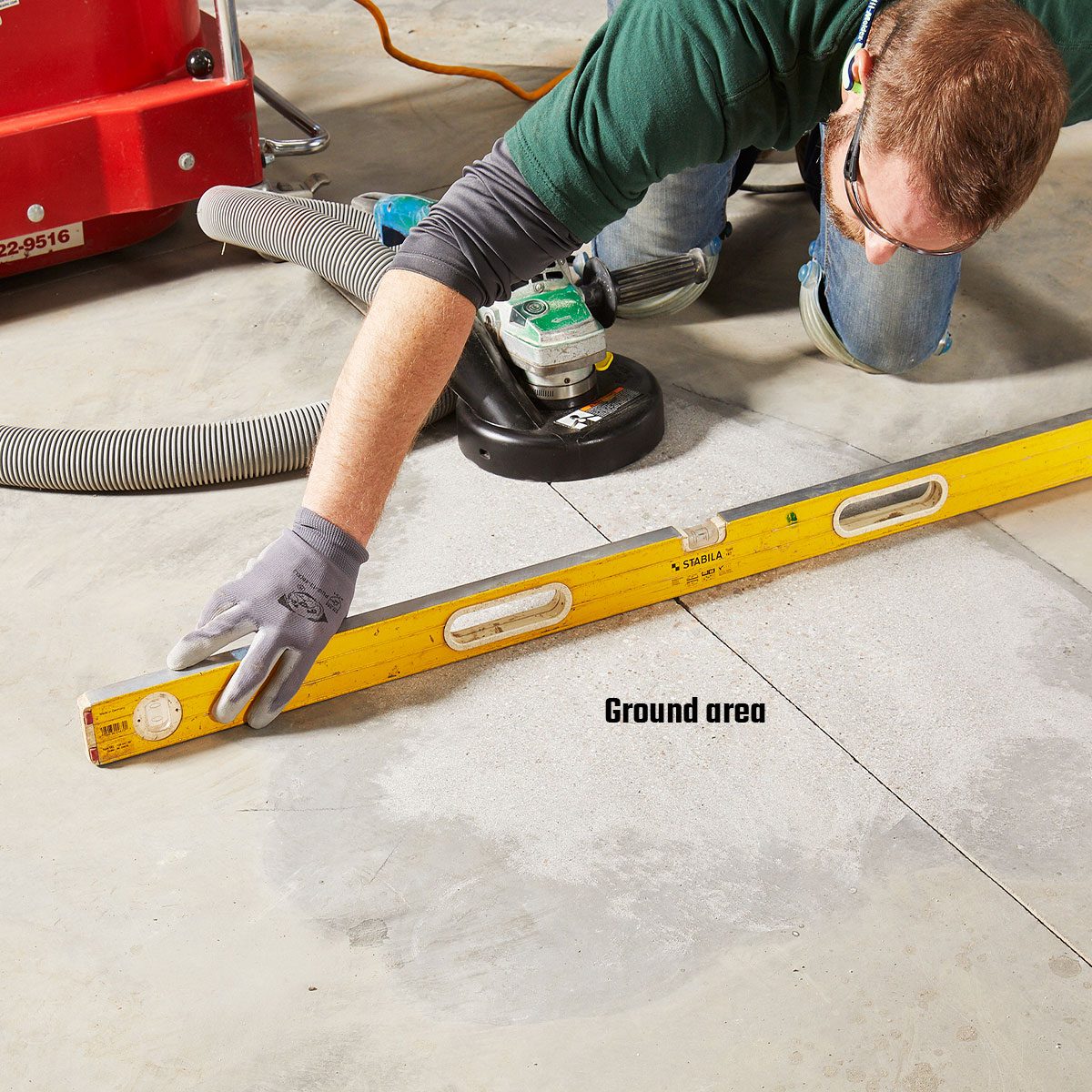 A person uses a level to check the flatness of a concrete floor, while equipment and a vacuum hose are nearby in a workspace.