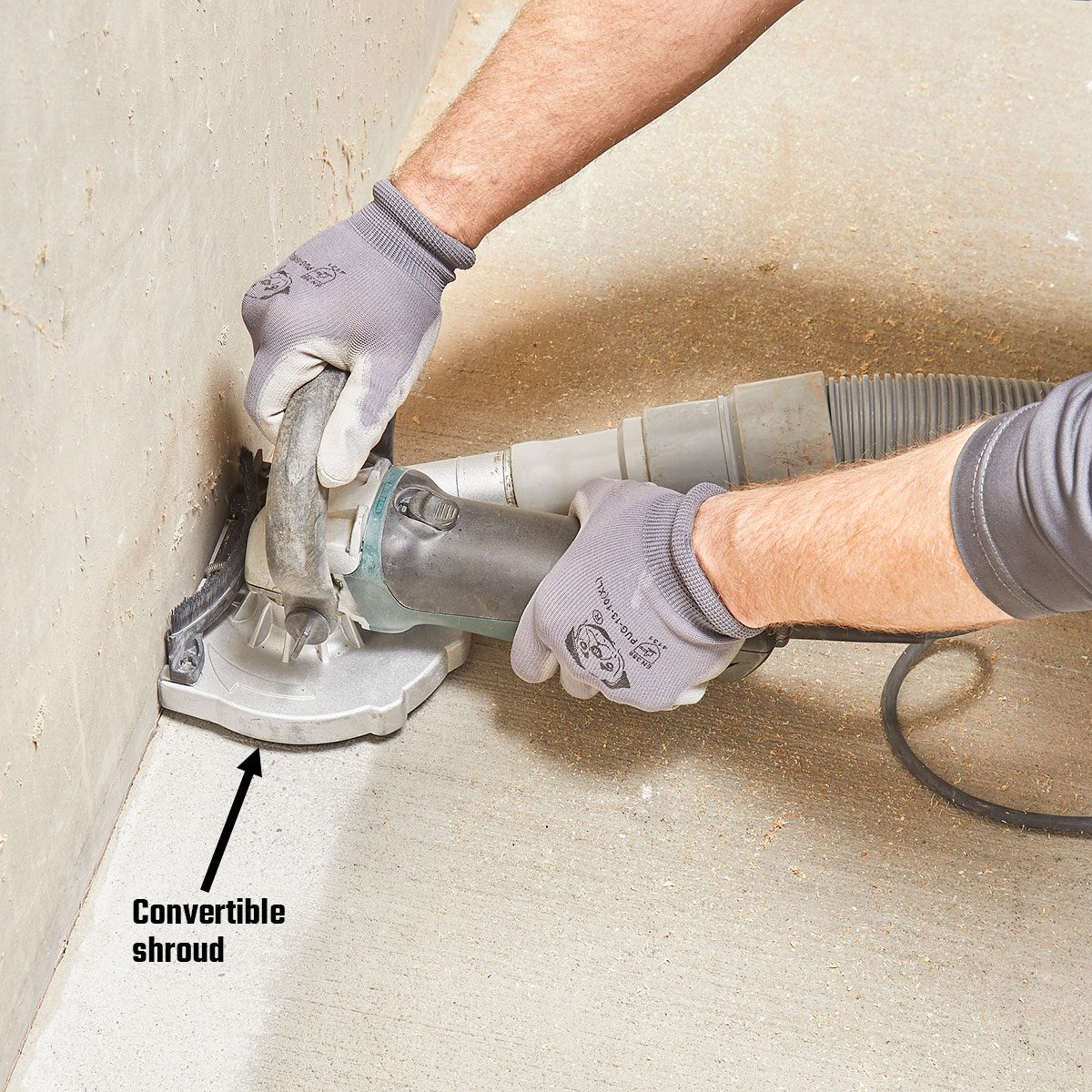 A person operates a power tool, adjusting a convertible shroud against a concrete wall, while wearing gloves, in a workshop environment.
