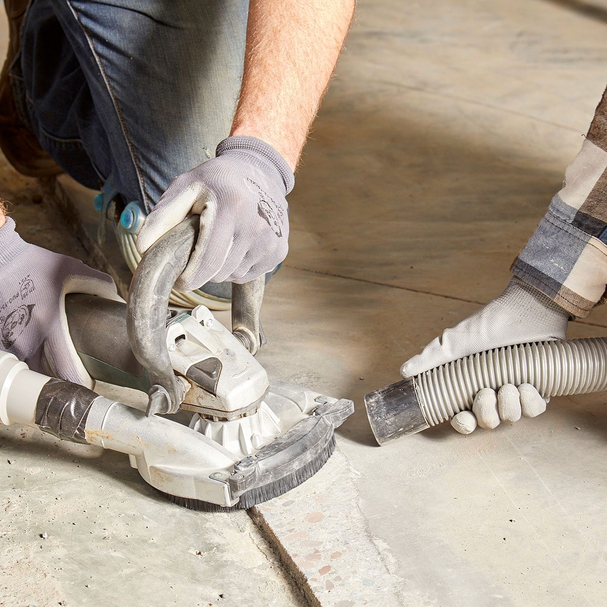 A person holds a power tool against concrete while another uses a vacuum hose, clearing dust in a well-lit indoor workspace.