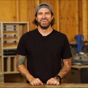 A man with tattoos stands smiling at a workbench, surrounded by wooden walls and tools, creating a welcoming atmosphere.