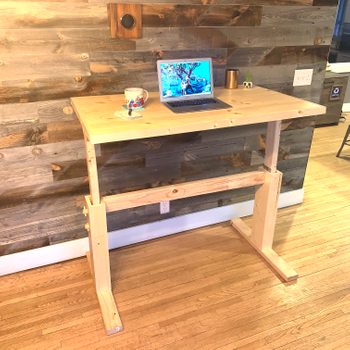 A wooden standing desk supports a laptop displaying a photo, with a mug and notebook nearby, set against a rustic wood-paneled wall.