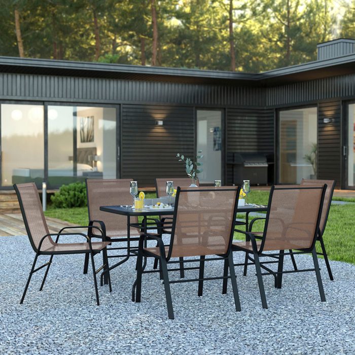 A glass-top table with six chairs is set with drinks and garnishes in a stone patio, surrounded by a modern house and greenery.