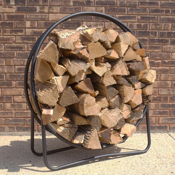 A circular wooden log holder displays neatly stacked firewood, resting on a black metal base against a textured brick wall.