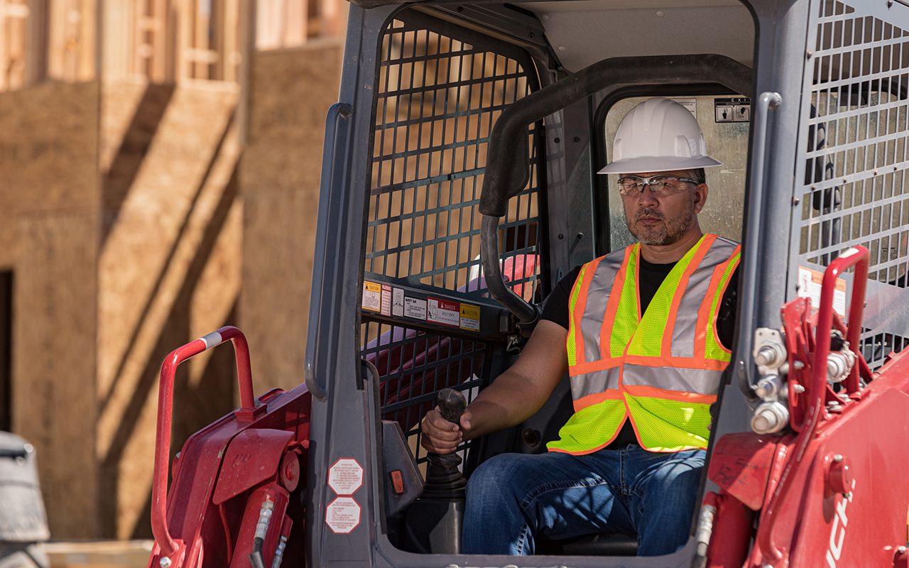 A man operates heavy machinery, focused within the cabin. Construction elements are visible in the background, indicating an active building site.