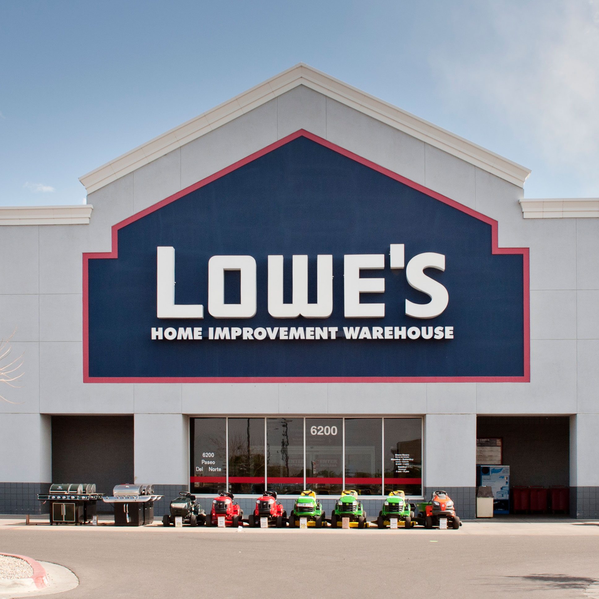 A Lowe's home improvement store features a large blue and red sign, displaying lawn mowers in front on a sunny day, with a clear blue sky above.