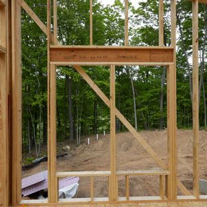 Wooden framing stands with cross-bracing, supporting a structure, overlooking a cleared area with soil and utility posts, surrounded by green trees.