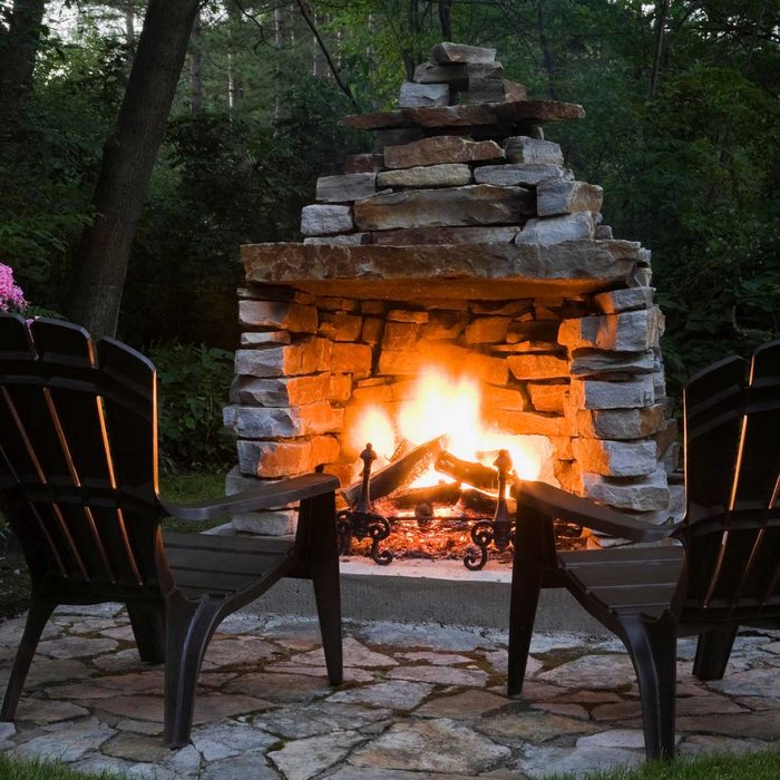 A stone fireplace emits a warm fire. Two black chairs are positioned nearby on a stone patio, surrounded by lush greenery in a tranquil outdoor setting.