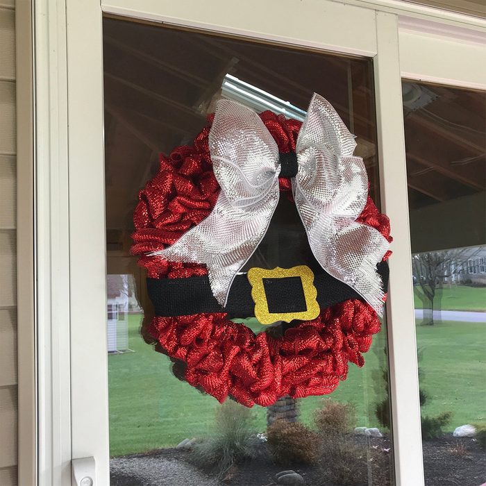 A decorative wreath resembling Santa's belt hangs on a door, featuring red and silver ribbons, a black belt, and a gold buckle, set in a yard.