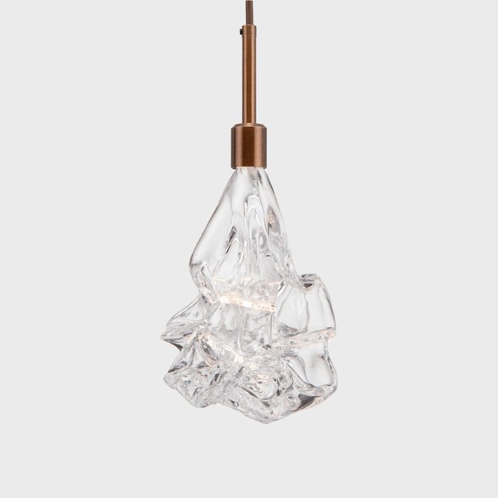 A clear, sculptural pendant light hangs from a bronze fixture, illuminating its unique, wavy design against a plain gray background.