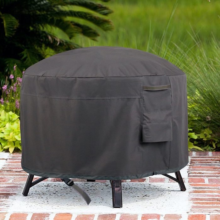 A round, dark cover with a pocket protects an outdoor table. It rests on a brick surface amidst green foliage and tall grass.