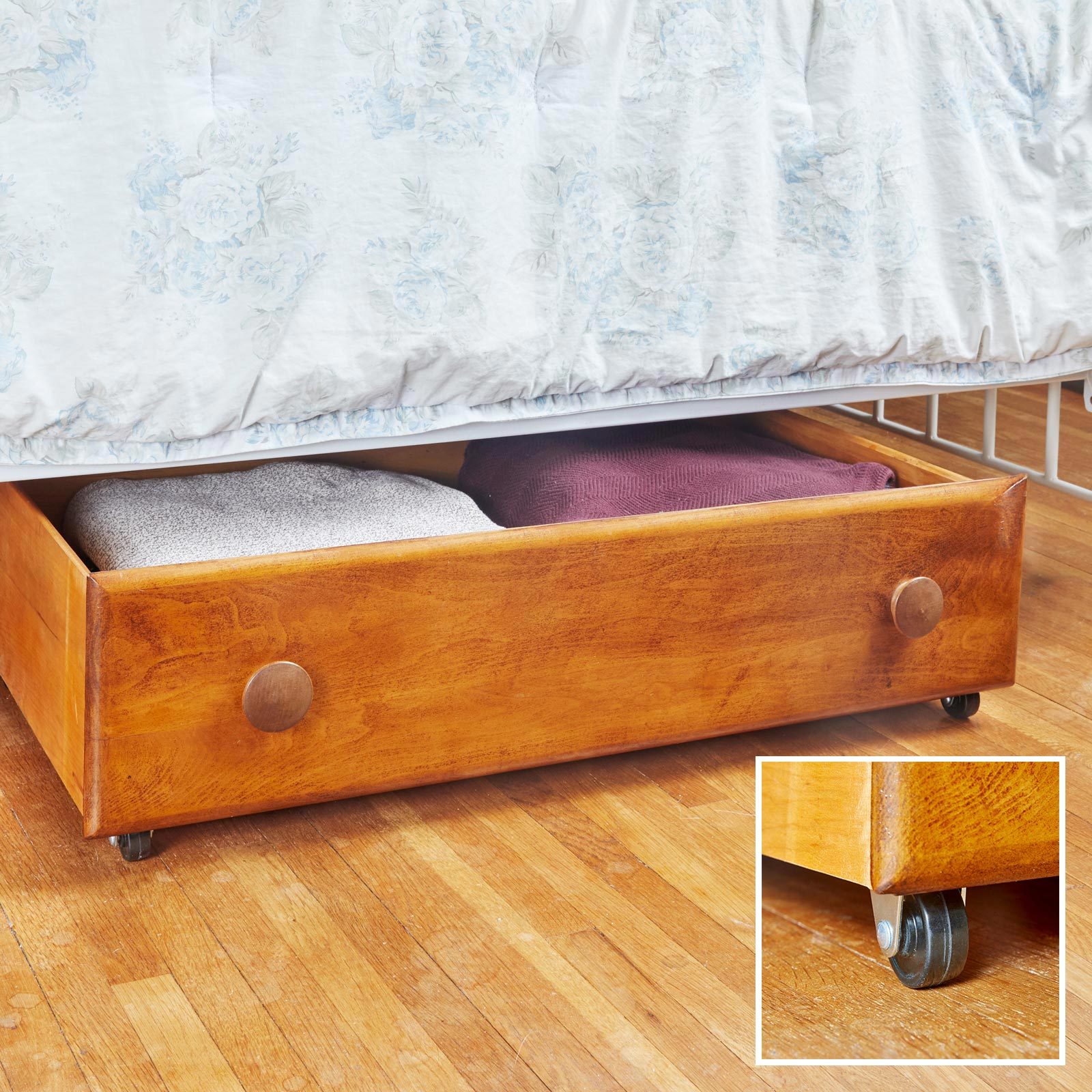 A wooden drawer, partially open, stores folded blankets. It is positioned underneath a bed, with floral bedding above and a wooden floor beneath.