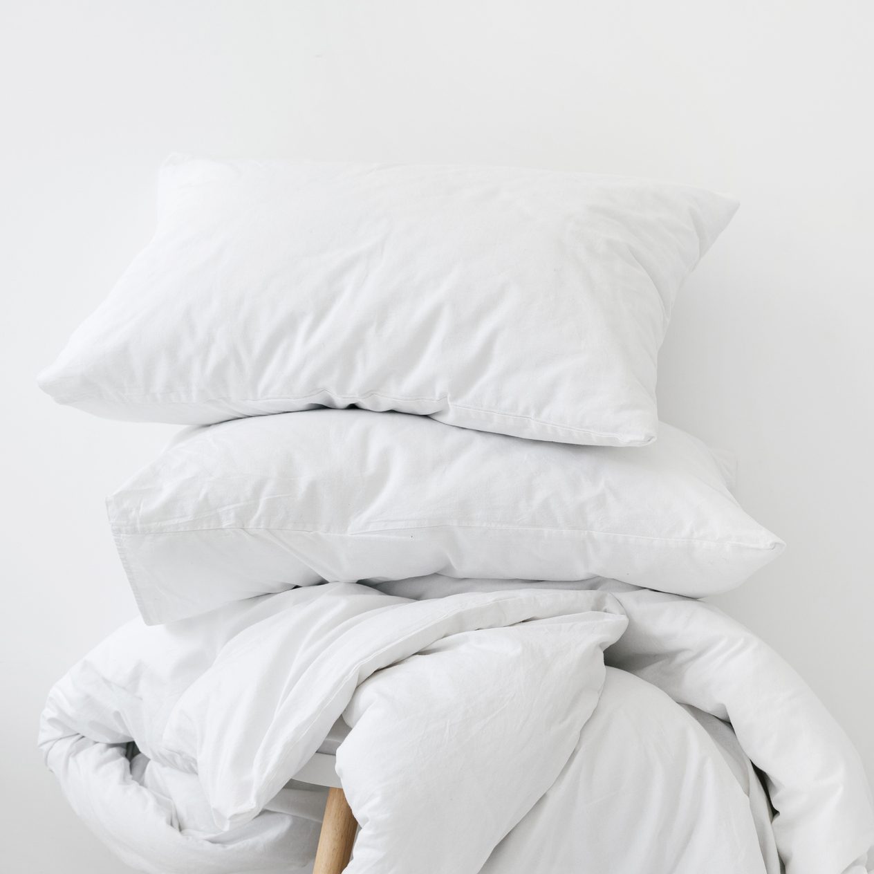 A stack of white pillows rests on a white blanket, both placed on a neutral, light-colored surface, creating a serene and minimalist bedroom atmosphere.