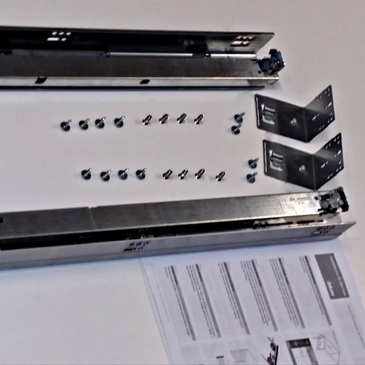 Two metal rails lie on a surface alongside screws and brackets, with an instruction sheet partially visible, suggesting an assembly process.