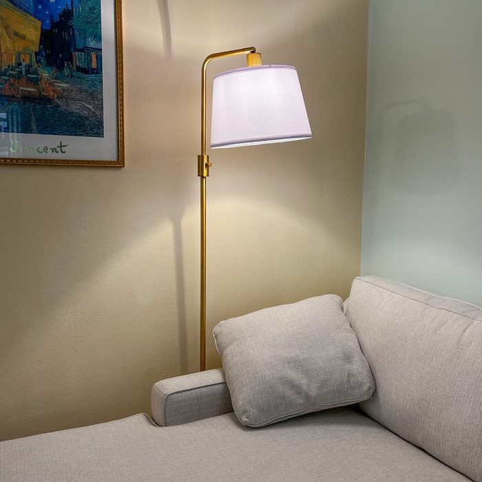 A gold floor lamp illuminates a light-colored couch, with a plush pillow resting against its arm, in a softly lit, cozy room.