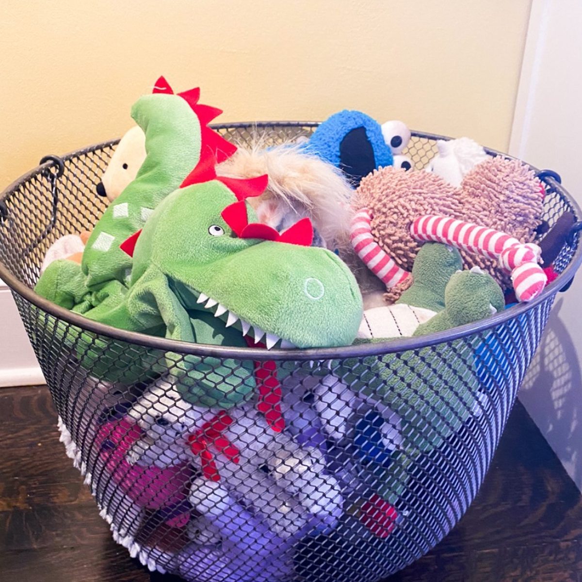 A wire basket contains various plush toys, including a green dinosaur, a teddy bear, and a cookie monster, arranged haphazardly against a light-colored wall.