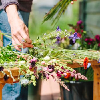 A hand arranges colorful flowers on a wooden table, surrounded by more blooms and greenery in a bright outdoor setting.