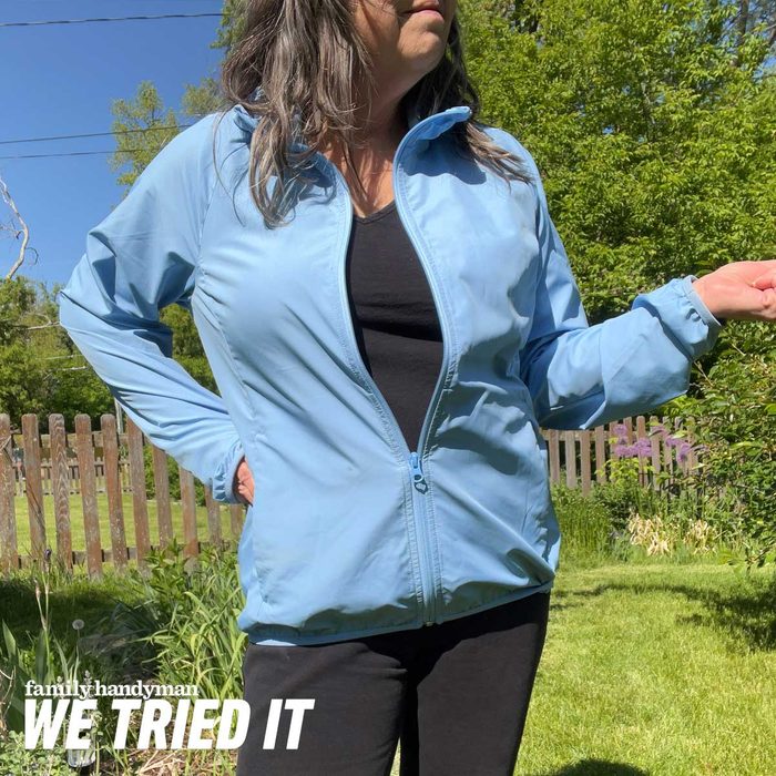 A woman in a blue jacket stands outdoors, gesturing with one hand while surrounded by green grass and trees under a clear blue sky.