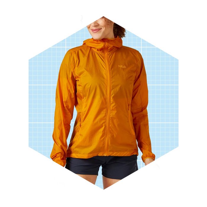 A woman wears an orange lightweight jacket with a hood, smiling against a light blue grid background.