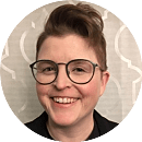 A person with short, styled hair and glasses smiles, against a light, patterned background, conveying a friendly and approachable demeanor.